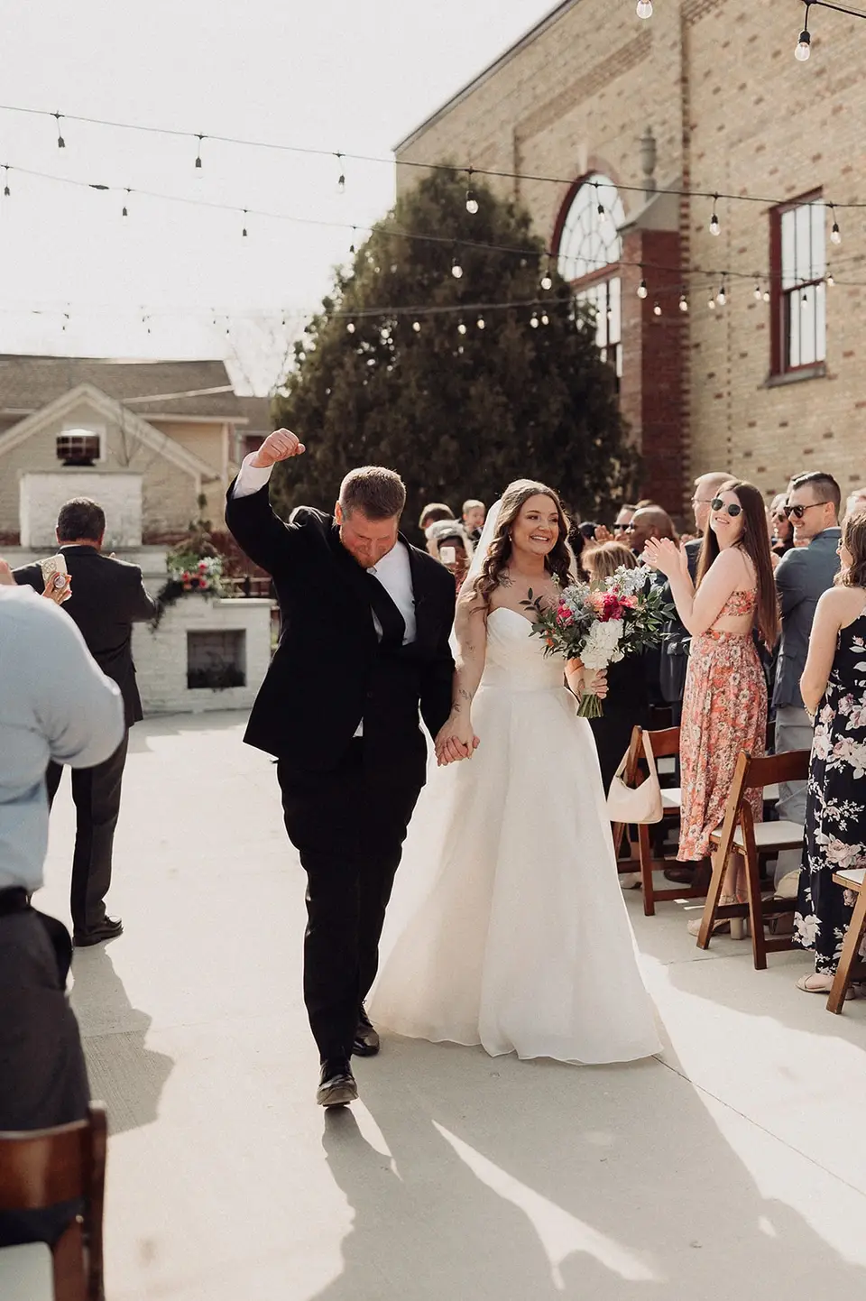 Just-married couple exits outdoor ceremony space at 10 South wedding venue in Janesville, WI, under string lights