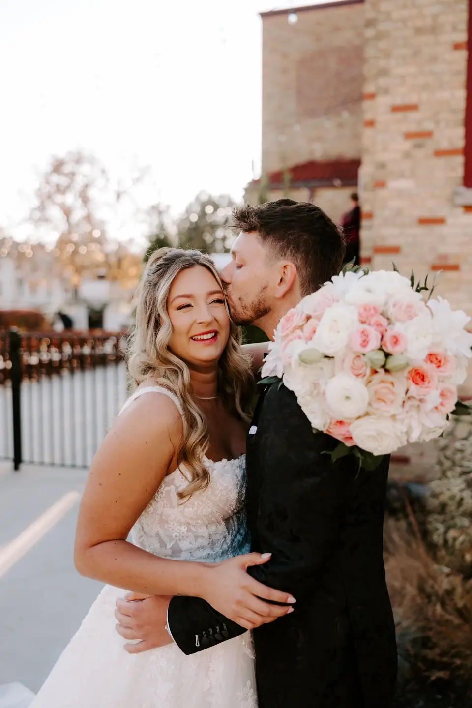 Groom kisses smiling bride on the cheek while holding a bouquet of blush and white roses outside 10 South in Janesville, Wisconsin
