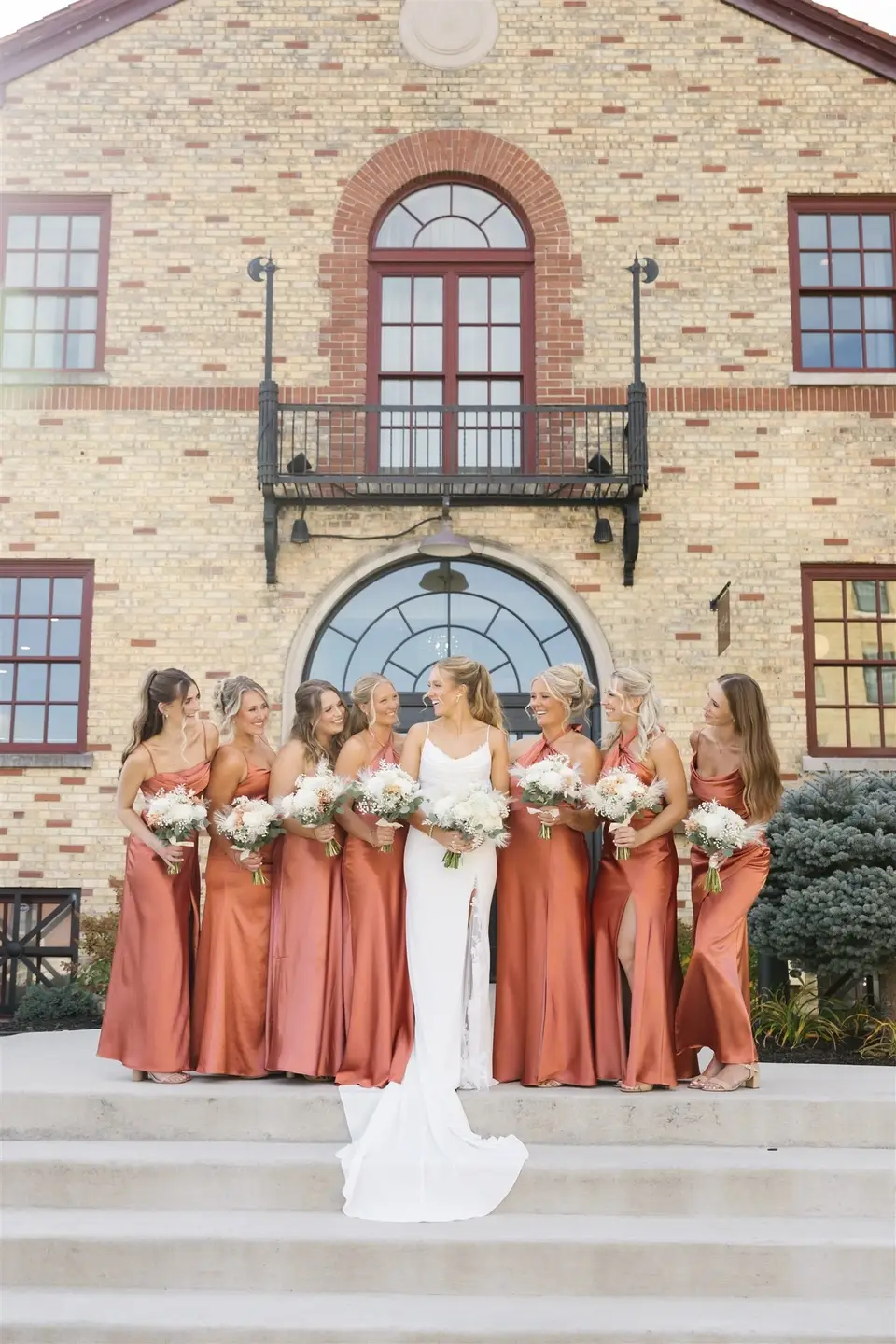 Bride and bridesmaids in rust satin dresses pose in front of 10 South wedding venue in Janesville, Wisconsin