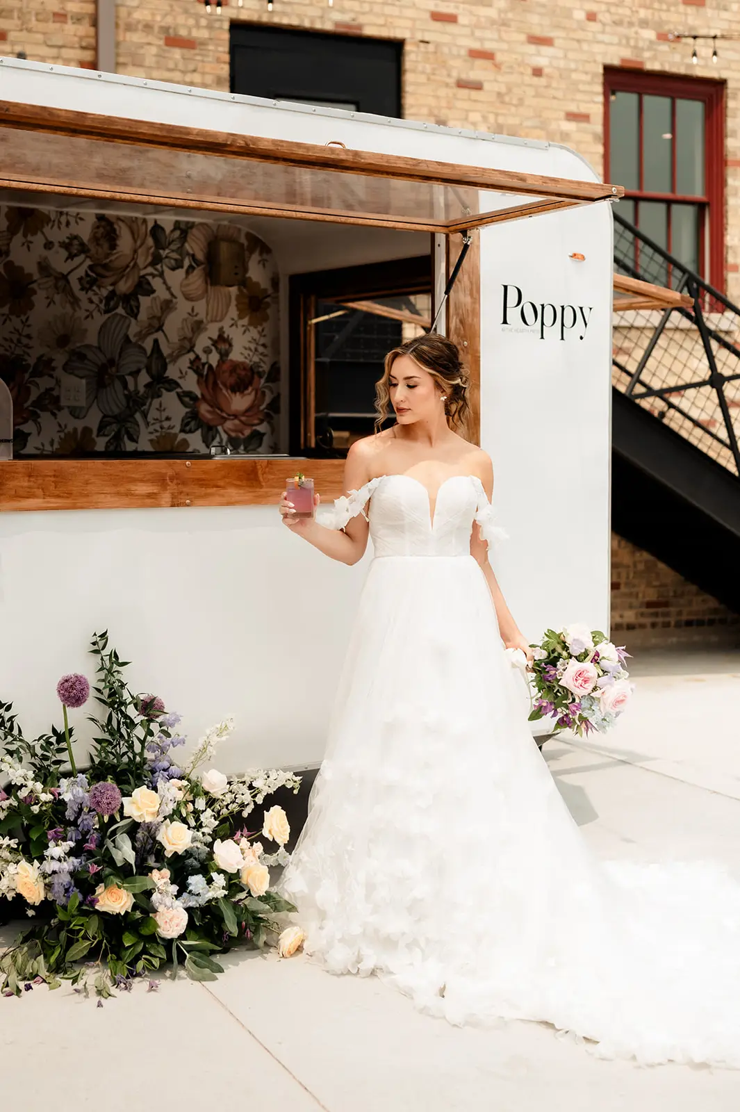 Bride in floral wedding gown stands beside Poppy, the vintage camper bar at 10 South in Janesville, Wisconsin