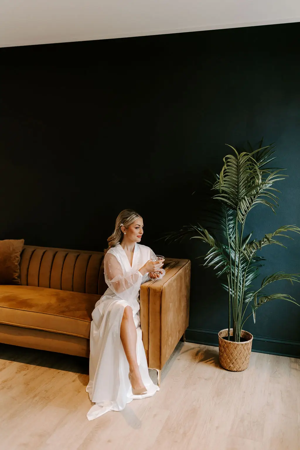 Bride in sheer-sleeved robe sits with a drink on a velvet amber couch in the modern lounge space at 10 South in Janesville, WI