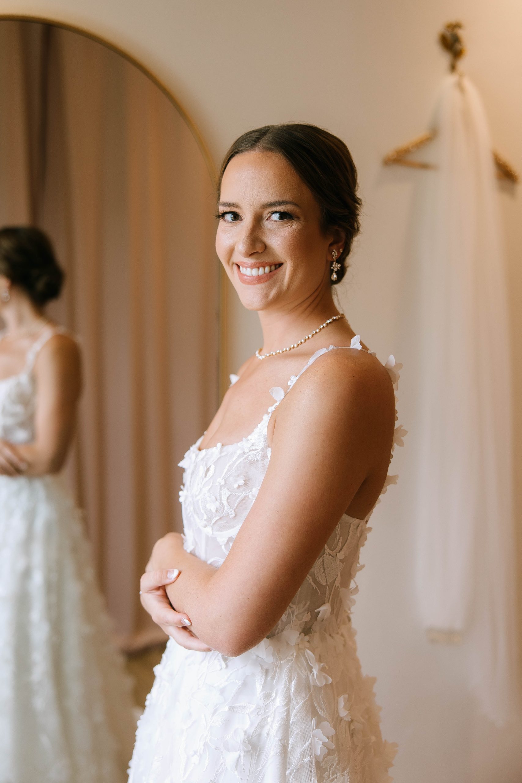 Bride smiles in a textured floral gown with pearl accessories, standing inside the bridal suite at 10 South.