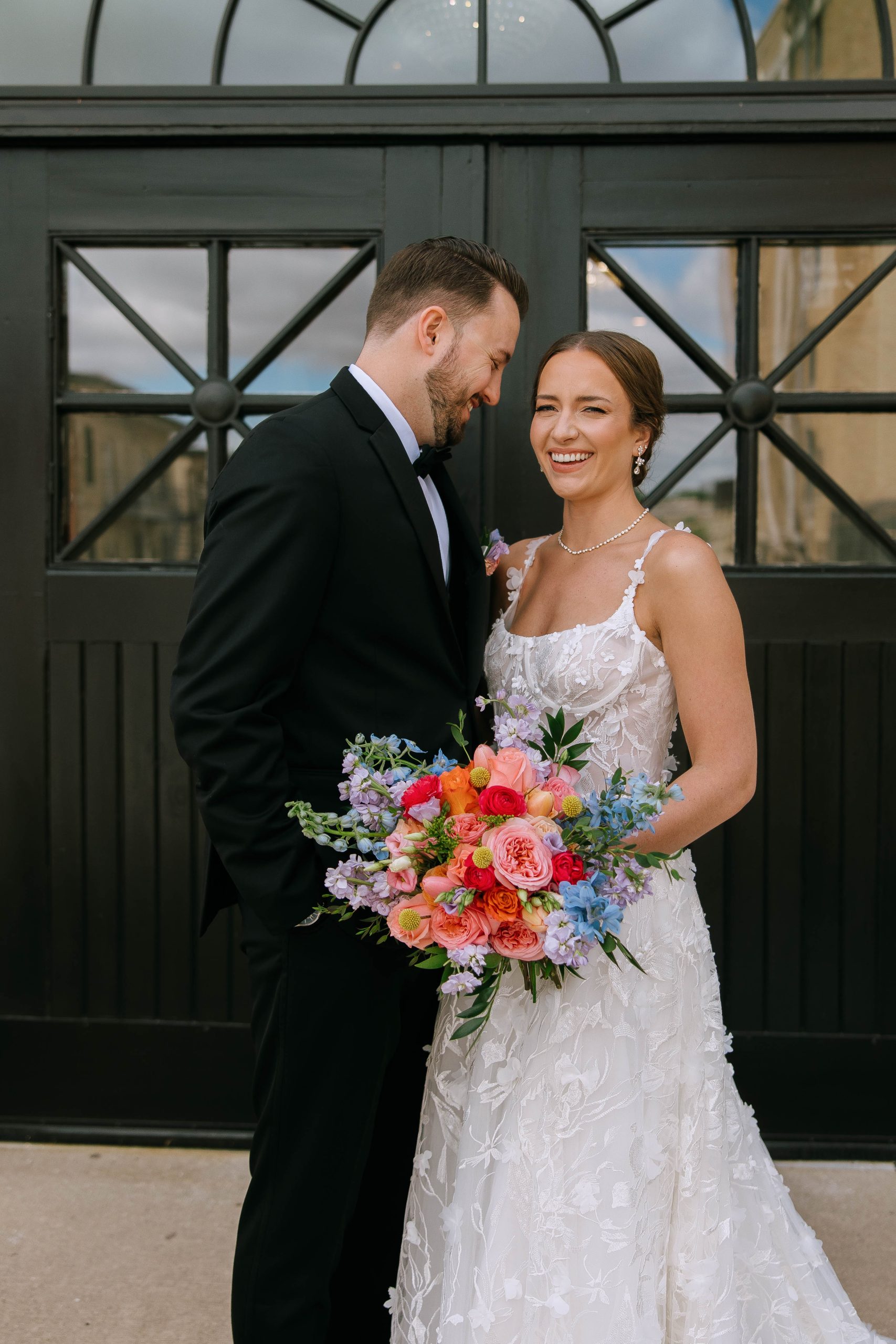 Bride and groom share a joyful moment in front of black arched doors at 10 South, holding a bright floral bouquet.