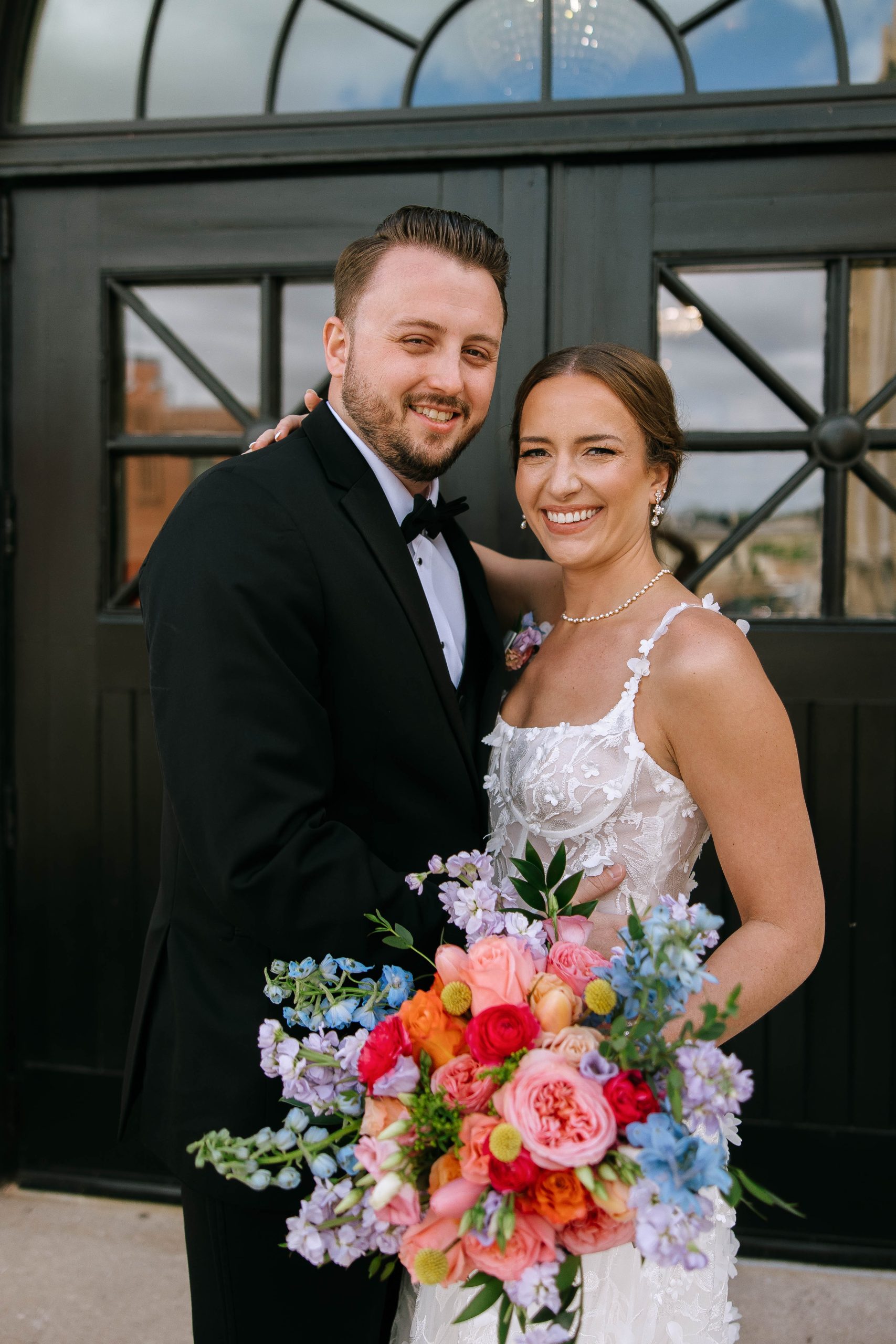 Bride and groom smiling in front of black arched doors at 10 South with a vibrant spring wedding bouquet.