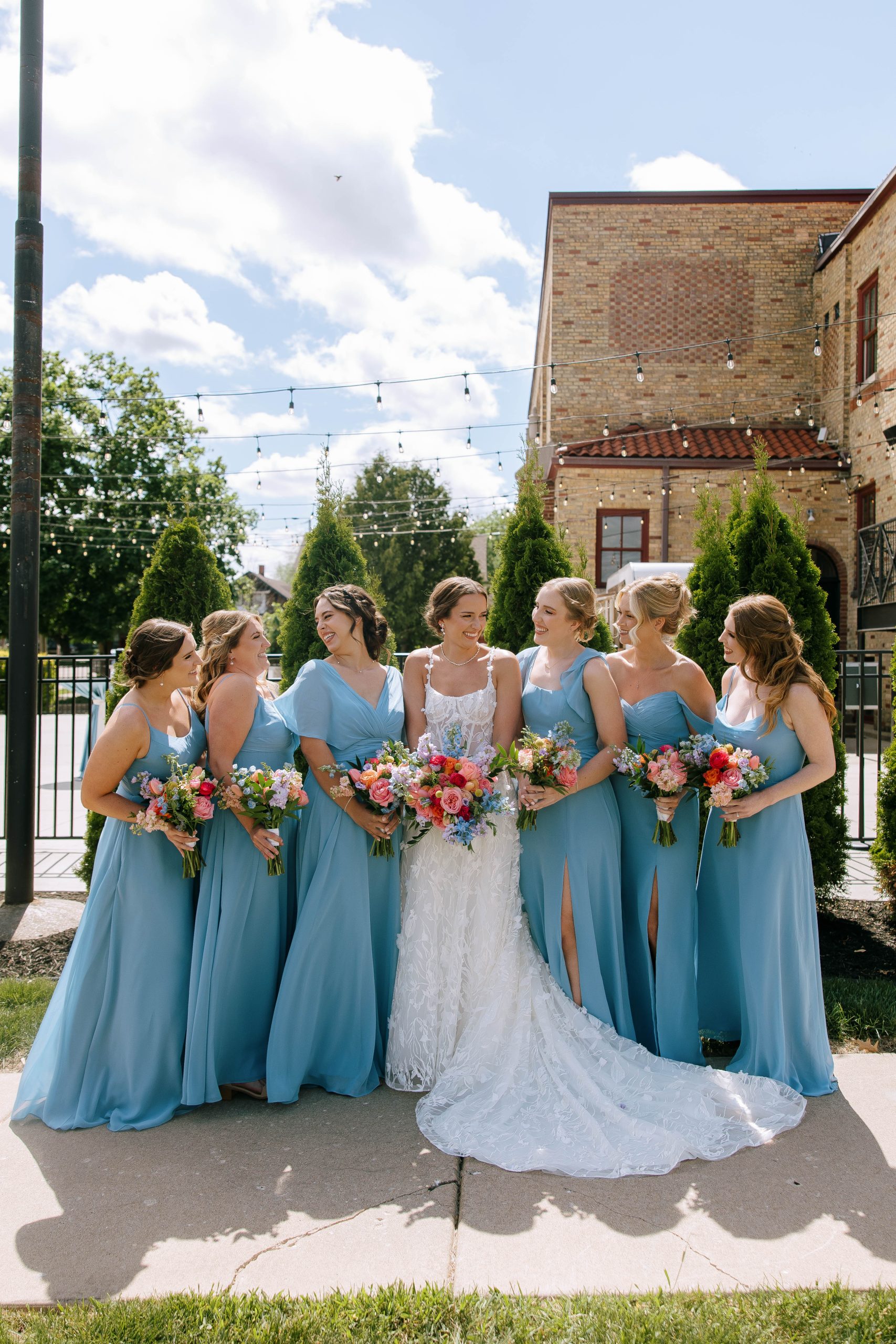 Bride stands with bridesmaids in matching blue gowns holding vibrant bouquets outside 10 South wedding venue.