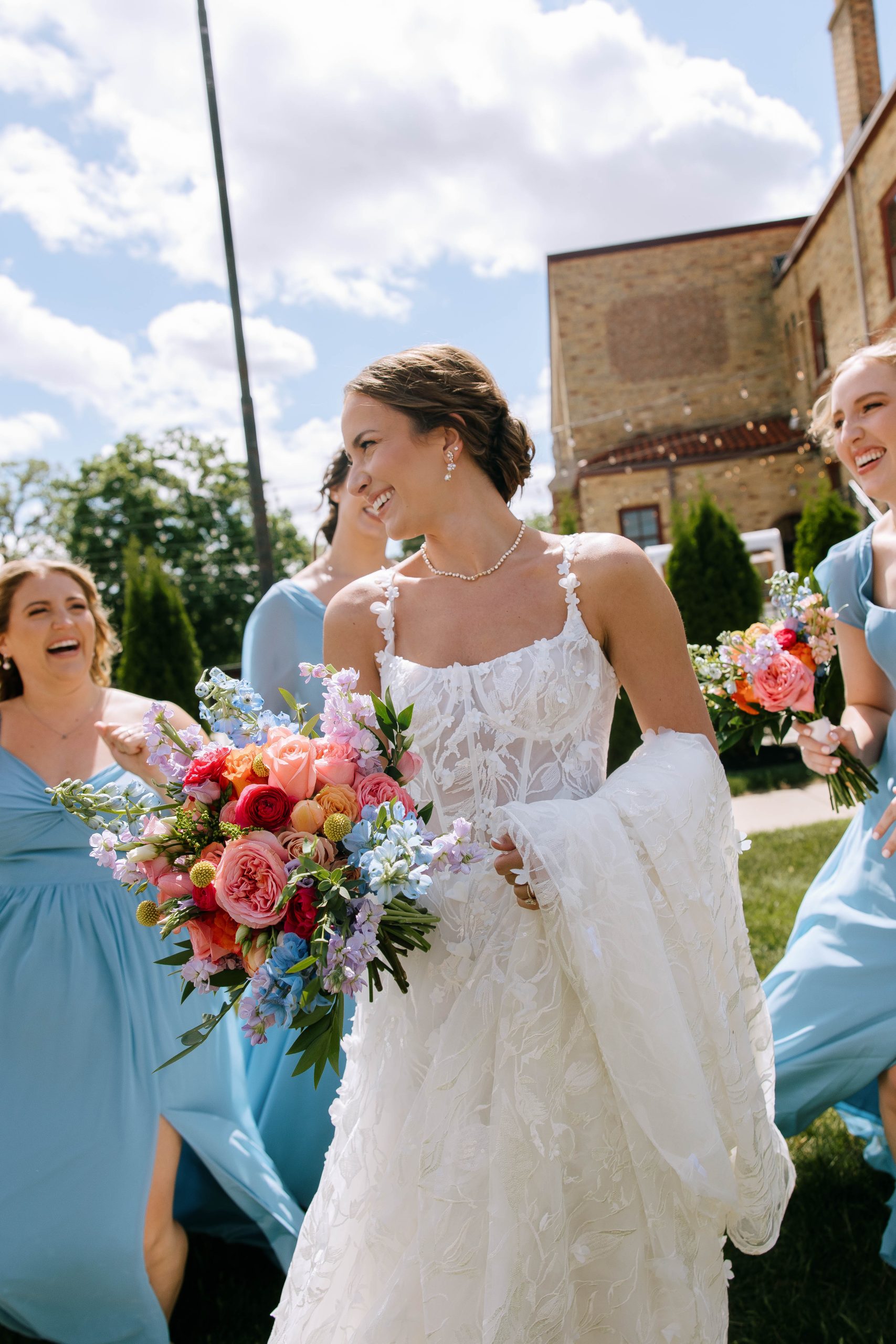 Bride laughing with bridesmaids in blue dresses on the lawn outside 10 South, holding a vibrant bouquet.