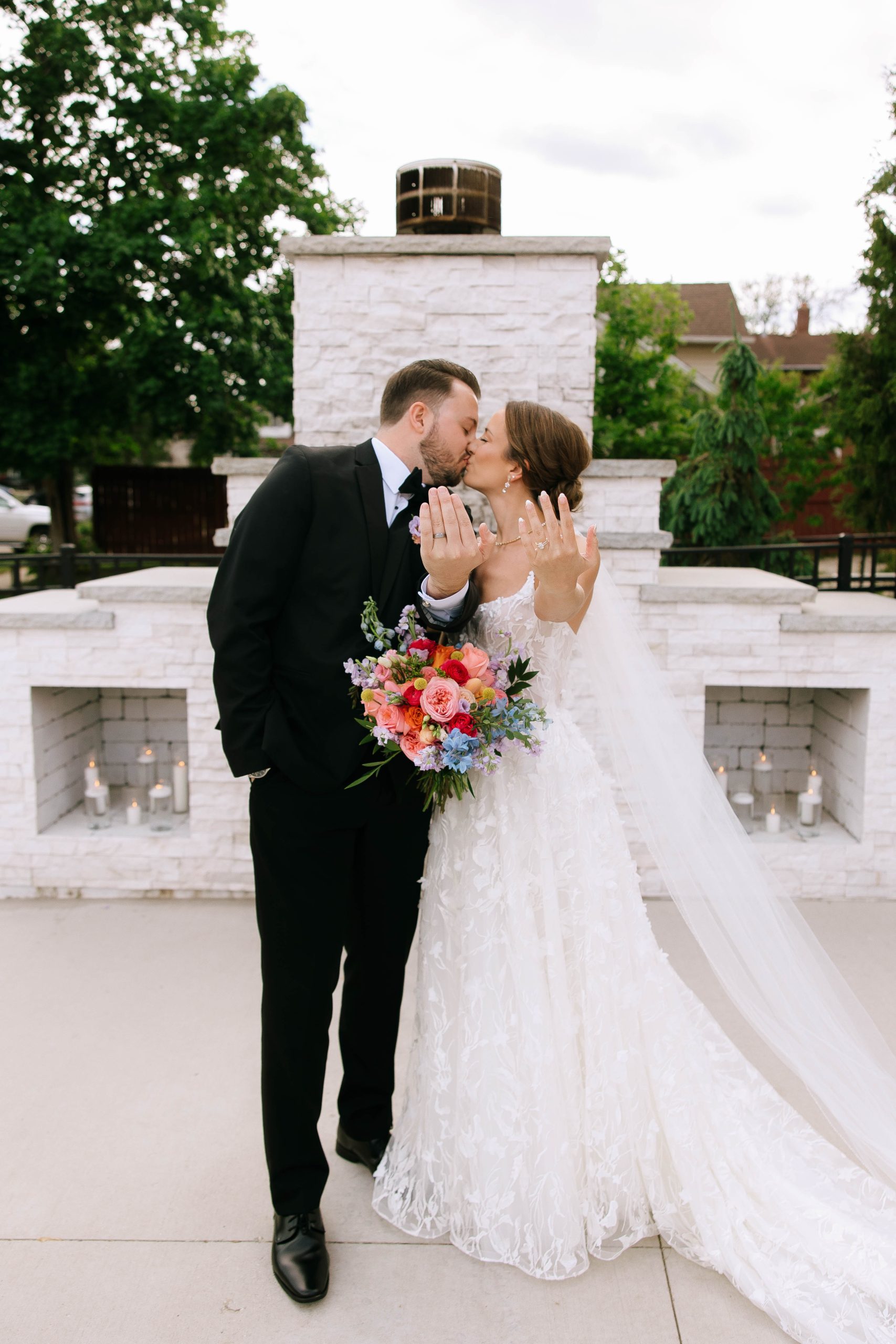 Bride and groom kiss while showing off their wedding rings in front of the outdoor fireplace at 10 South in Janesville, WI.