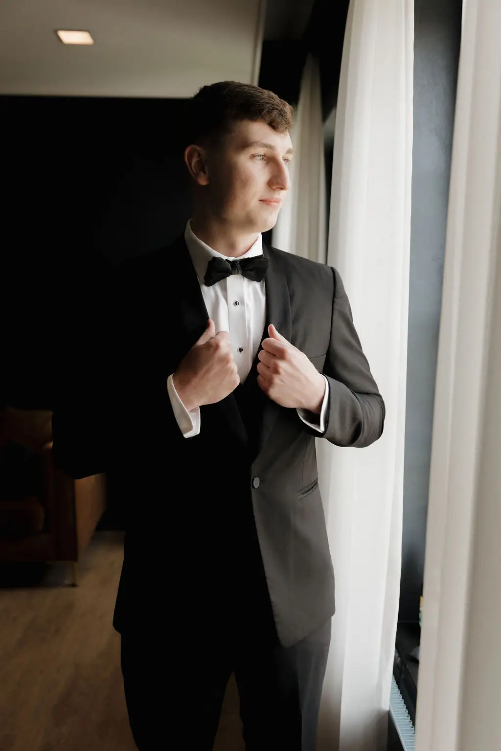 Groom in black tuxedo adjusts jacket while looking out the window in the groom’s lounge at 10 South in Janesville, WI