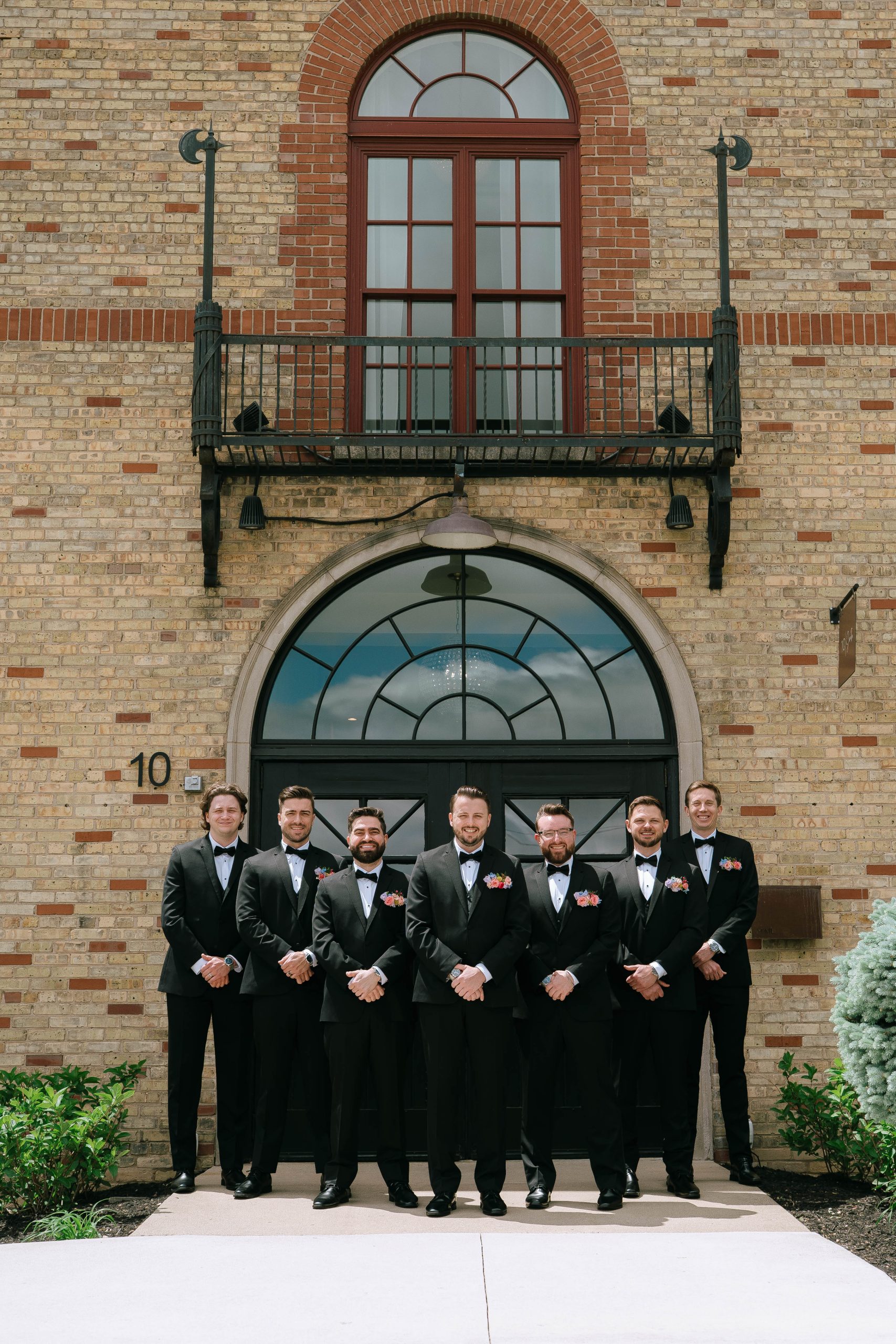 Groom and groomsmen in black tuxedos pose confidently outside the historic brick facade of 10 South wedding venue in Janesville, Wisconsin.