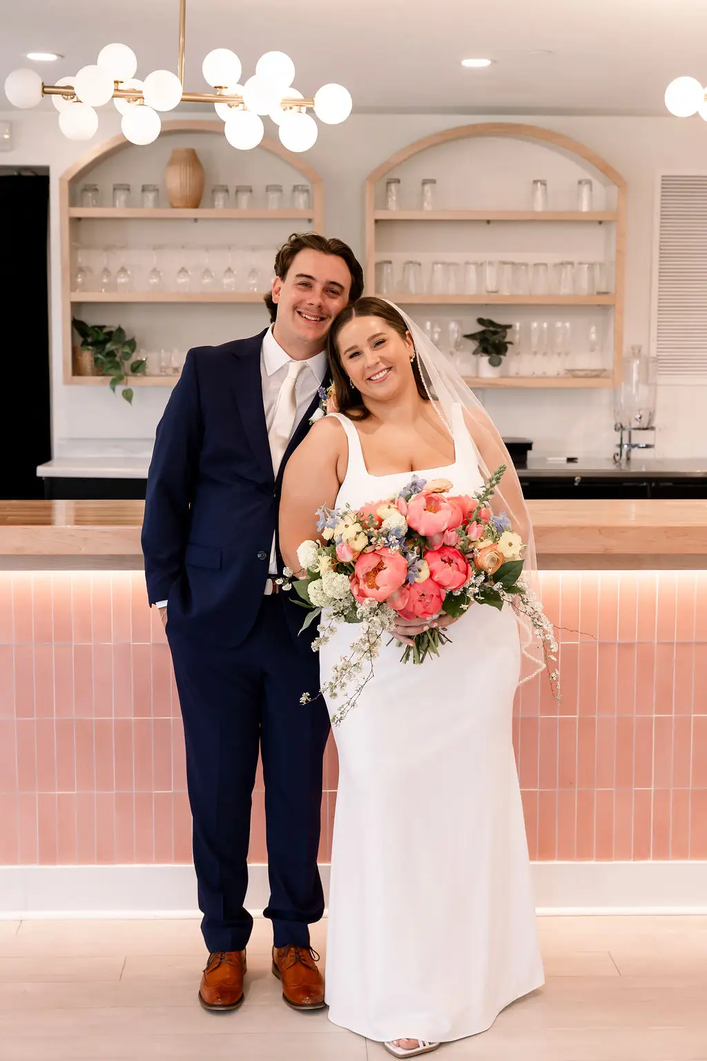 Bride and groom smile in front of modern pink tile bar with floral bouquet at 10 South wedding venue in Janesville, WI