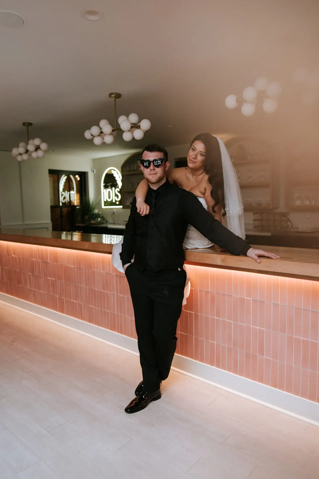 Bride poses with groom in all-black suit and sunglasses at the modern indoor bar at 10 South wedding venue in Janesville, WI