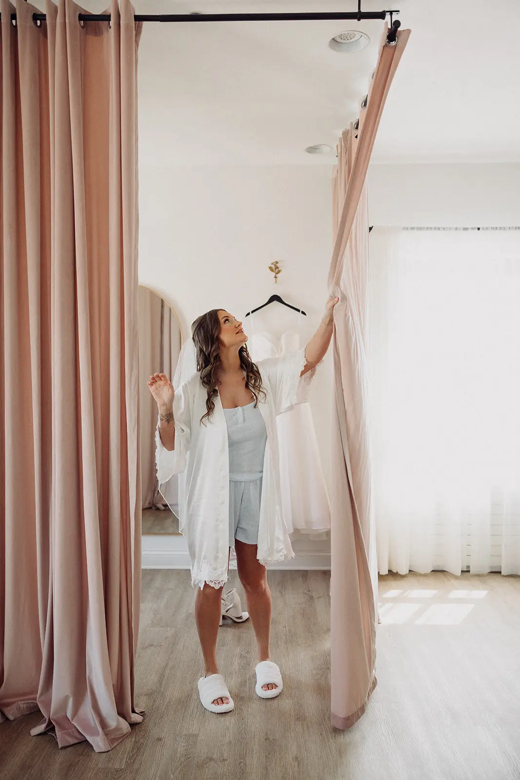 Bride in robe stands beside blush curtains with her wedding dress hanging behind her in 10 South’s bridal suite in Janesville, WI