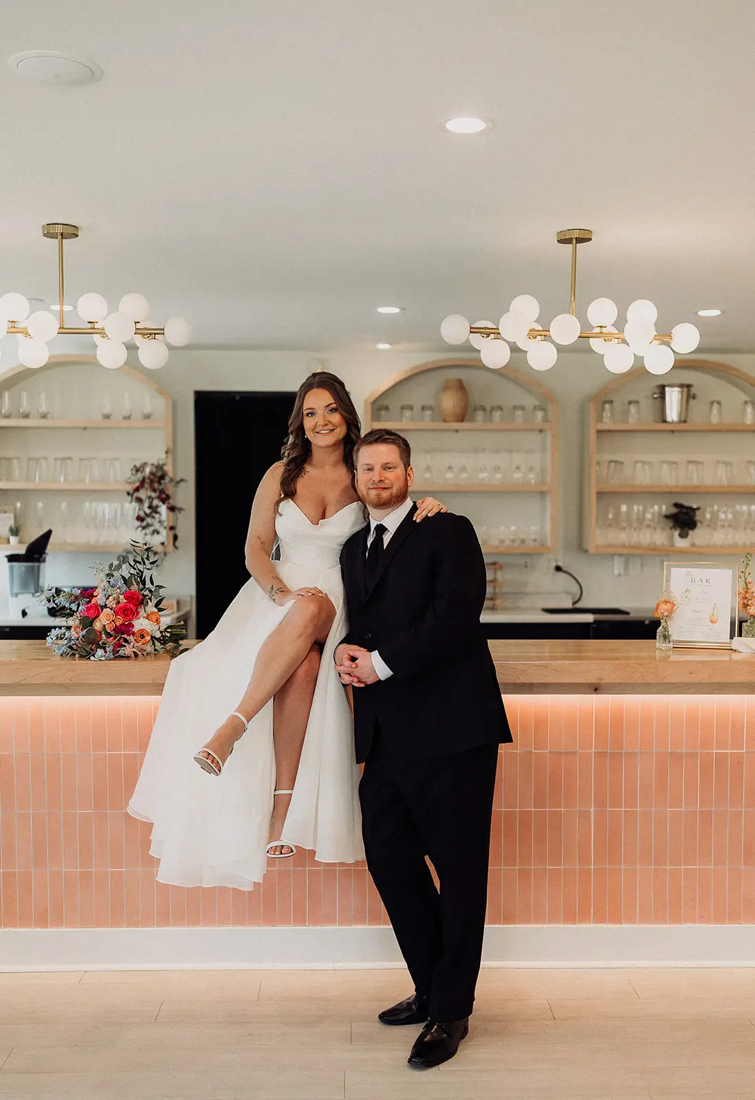 Bride sits on modern pink tile bar with groom standing beside her at 10 South wedding venue in Janesville, WI