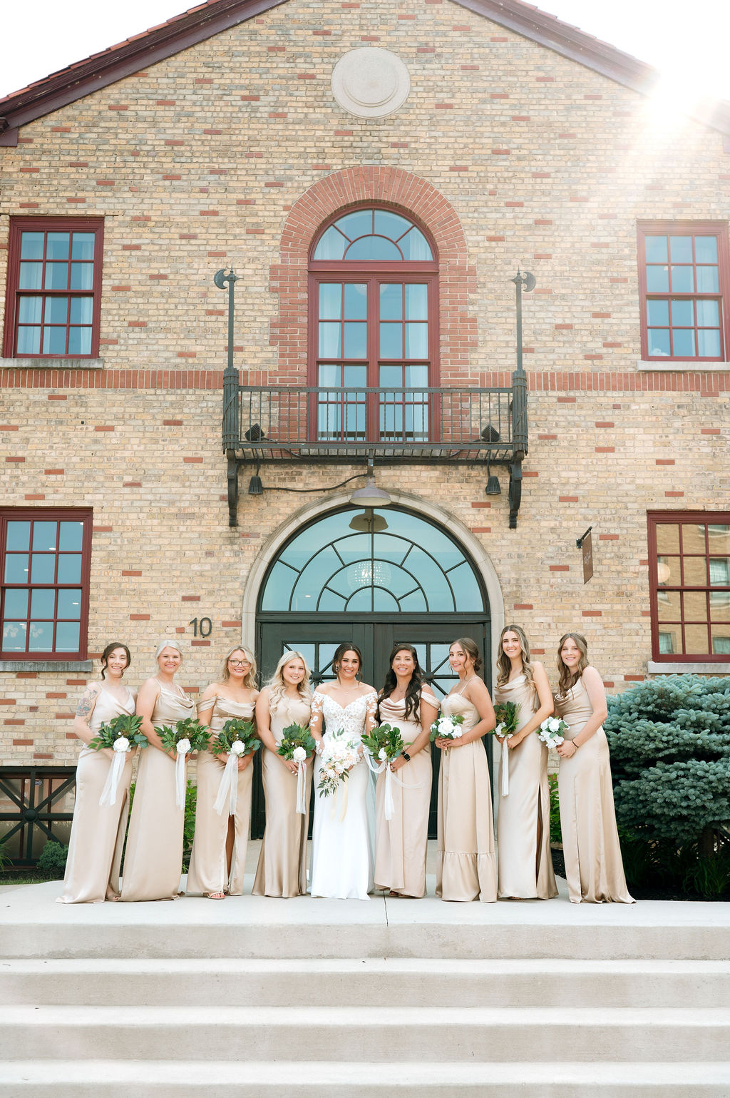 Bride and bridesmaids in champagne gowns stand on steps before 10 South’s brick facade.