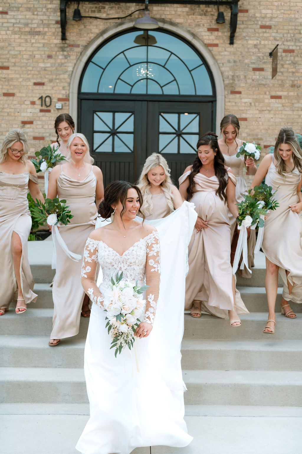 Bride in lace gown walks down venue steps with bridesmaids in champagne dresses at 10 South in Janesville, WI.