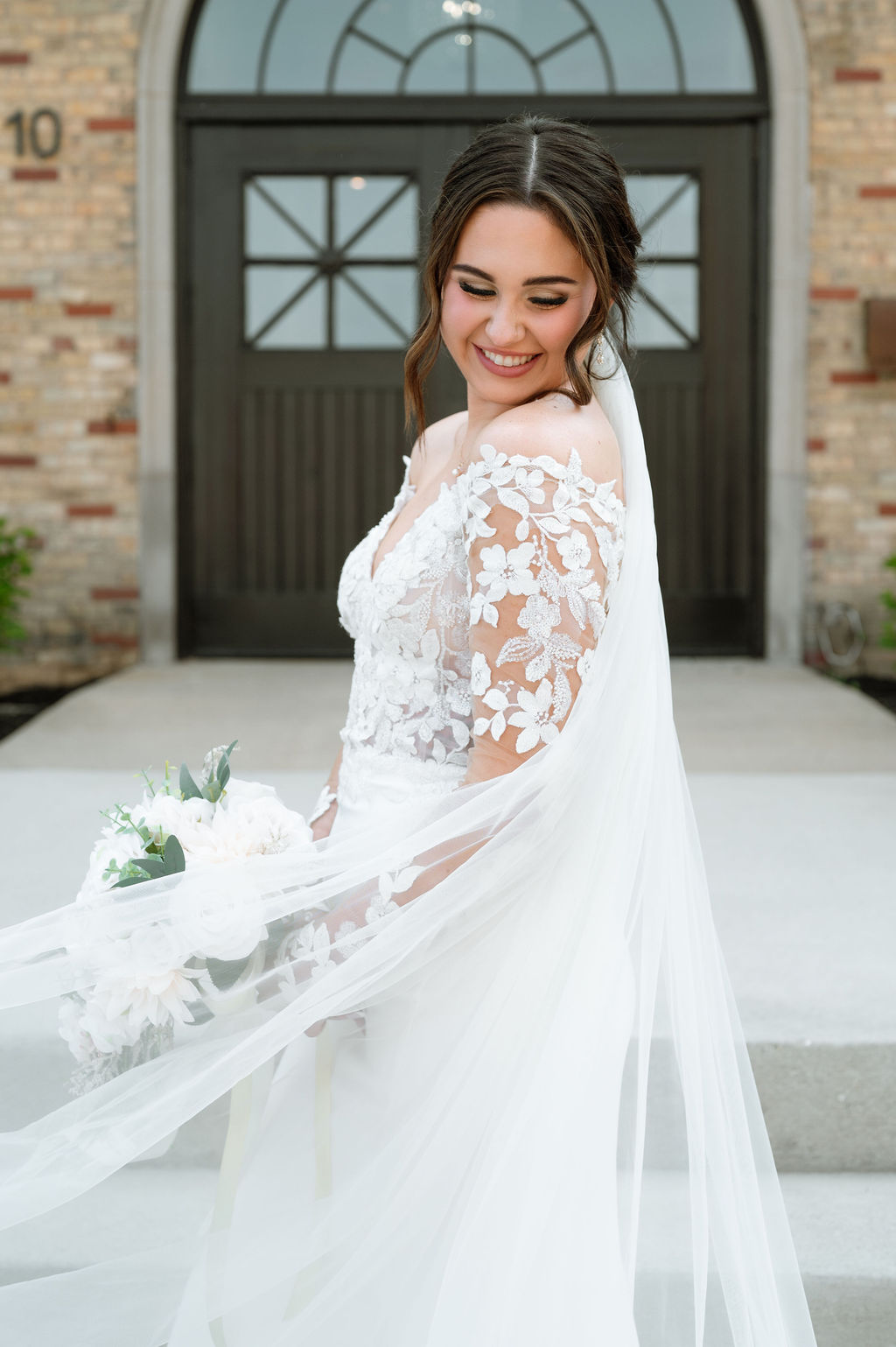 Bride in lace-sleeved gown and flowing veil smiles before 10 South’s arched doorway in Janesville.