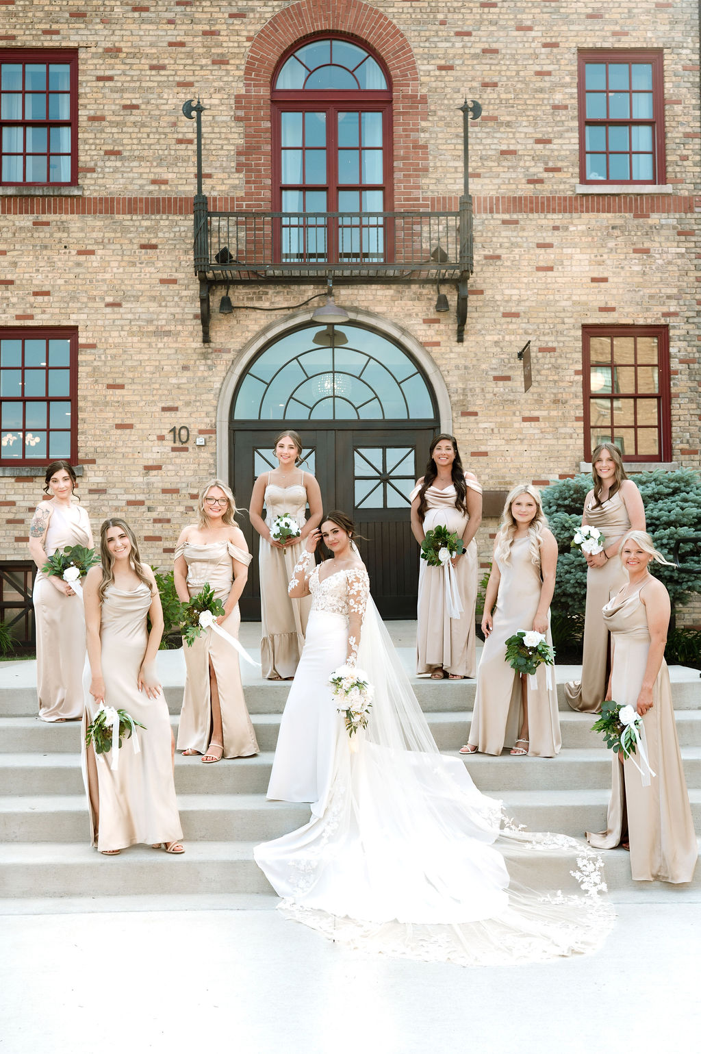 Bride in long train wedding dress poses with bridesmaids in champagne gowns on the front steps of 10 South wedding venue in Janesville, WI.