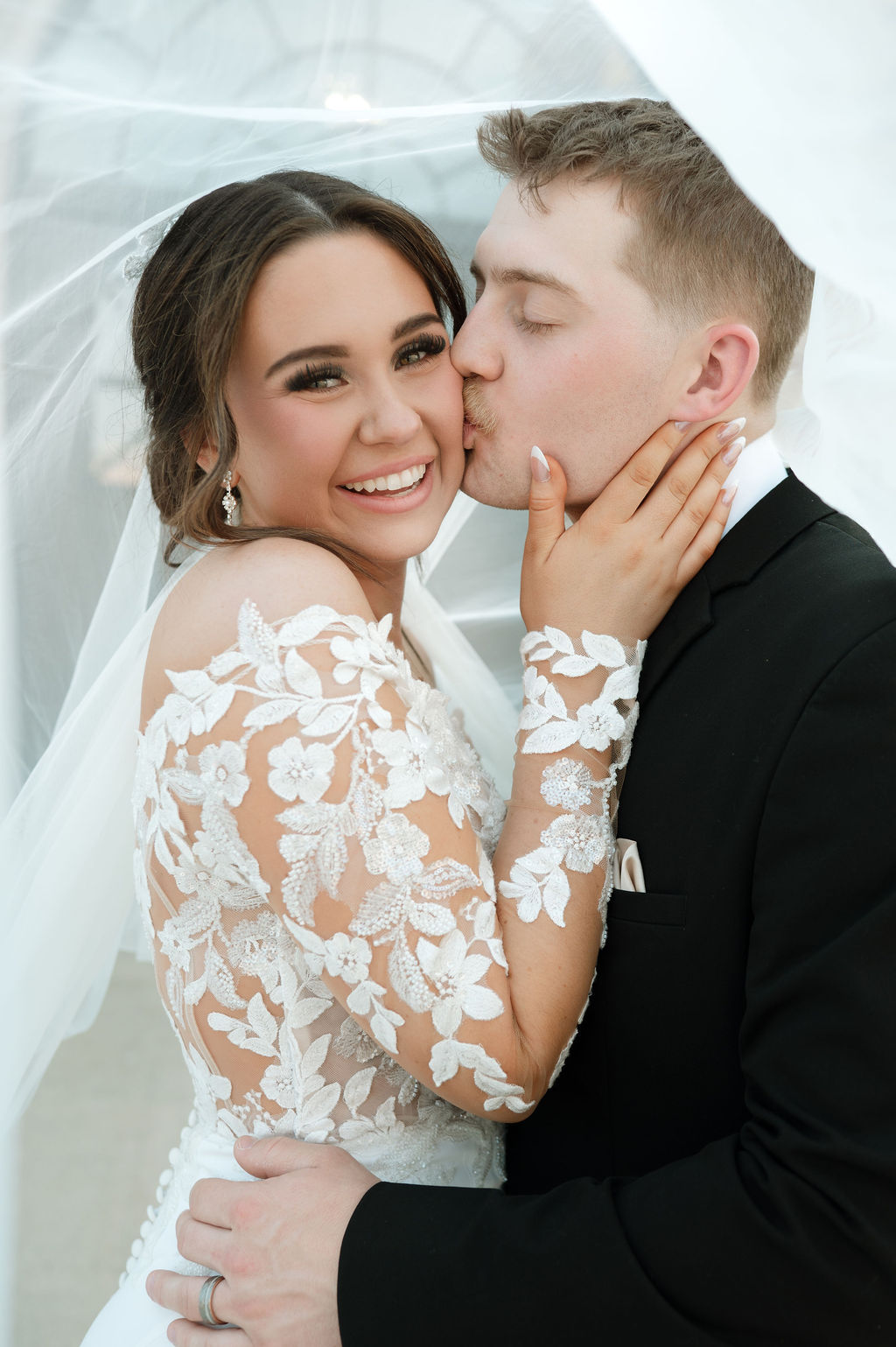 Groom tenderly kisses smiling bride on the cheek beneath her flowing veil in front of 10 South’s grand entrance.