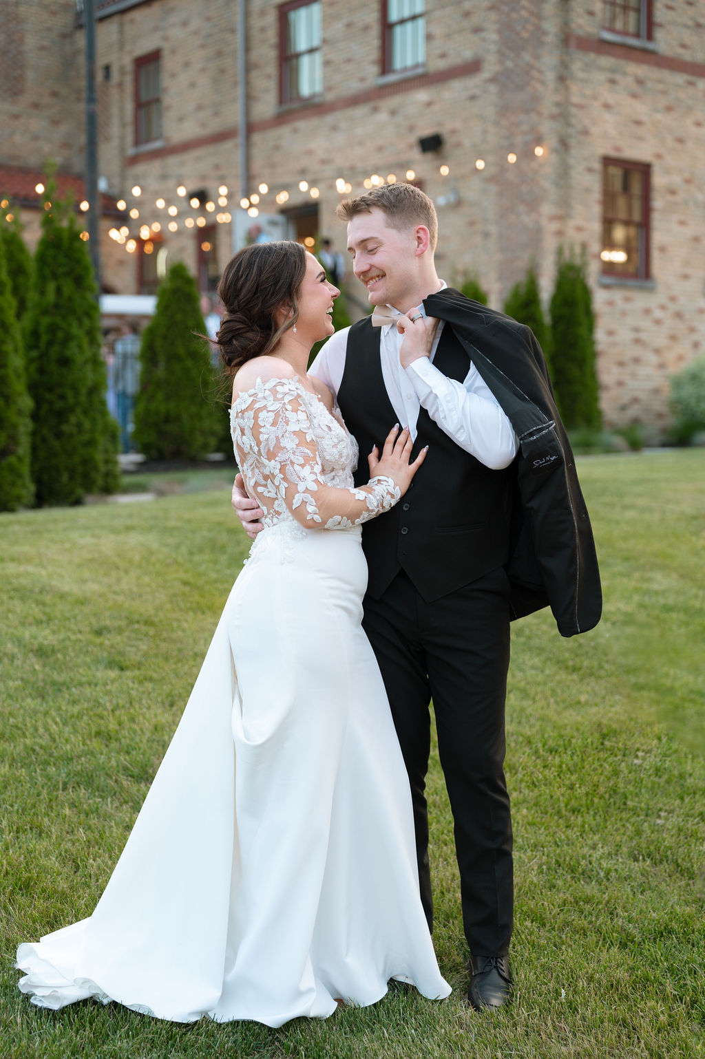 Bride and groom smiling at each other on the lawn at 10 South in Janesville, Wisconsin, with string lights and exposed brick building in the background.