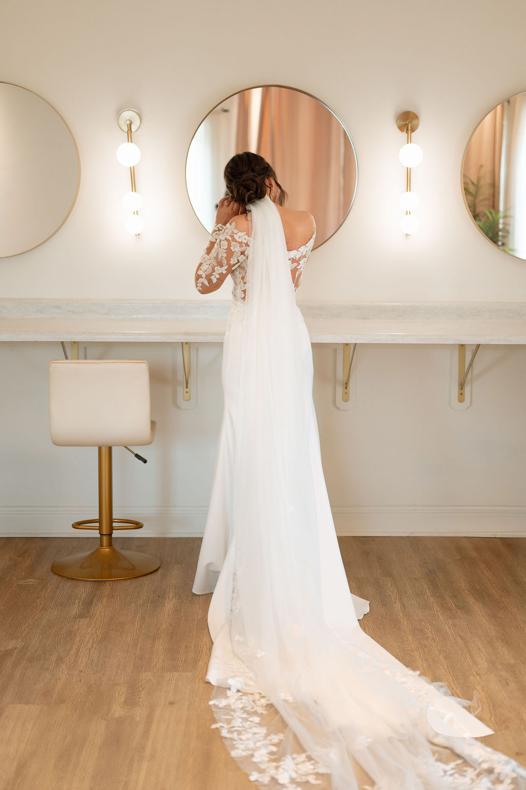 Bride in off-the-shoulder lace gown with chapel-length veil stands at the mirror in the bridal suite at 10 South in Janesville, WI.