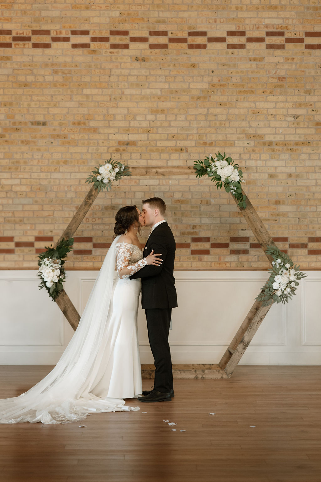 Bride and groom share a kiss in front of a wooden hexagon arch during their indoor wedding ceremony at 10 South in Janesville, WI.