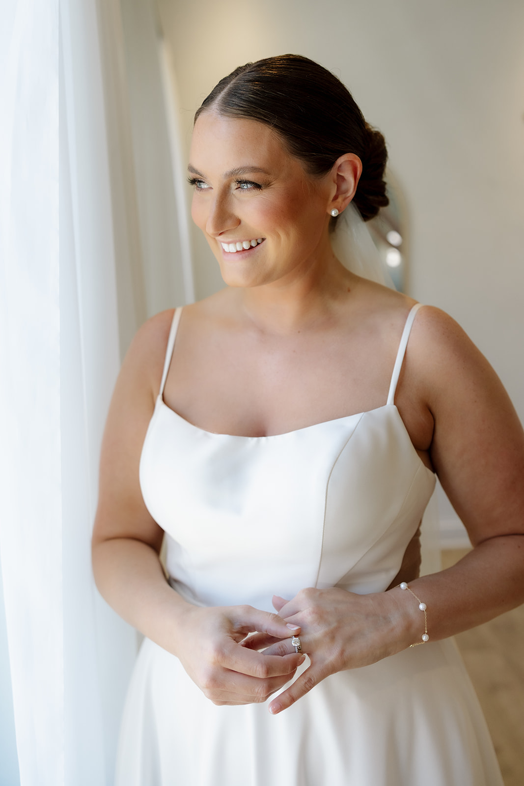 Bride in a modern satin gown smiles softly while standing beside a window at 10 South.