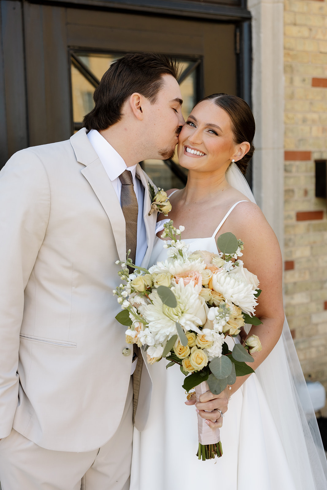 Groom kisses bride’s cheek as she smiles holding a soft ivory and peach bouquet outside 10 South.