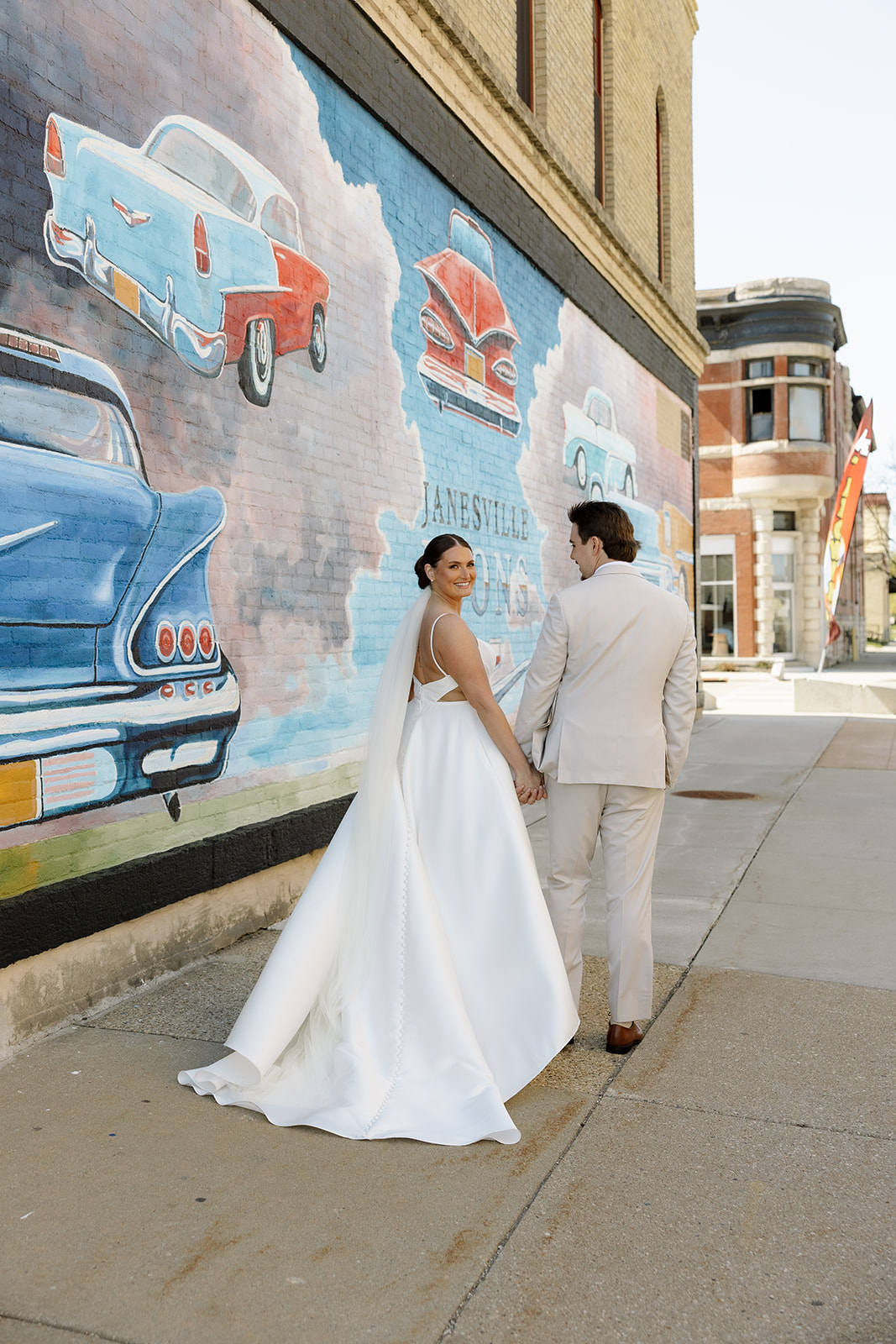 Bride and groom walk hand-in-hand past the colorful Janesville car mural on their wedding day.
