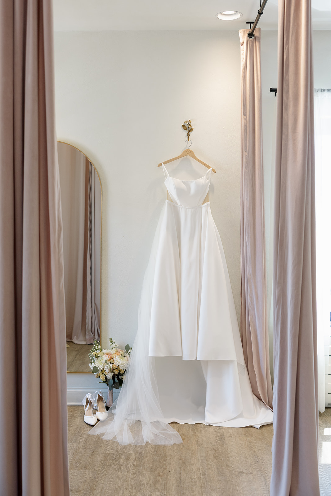 Modern ivory wedding dress hanging in a bridal suite with blush curtains and a bouquet below.