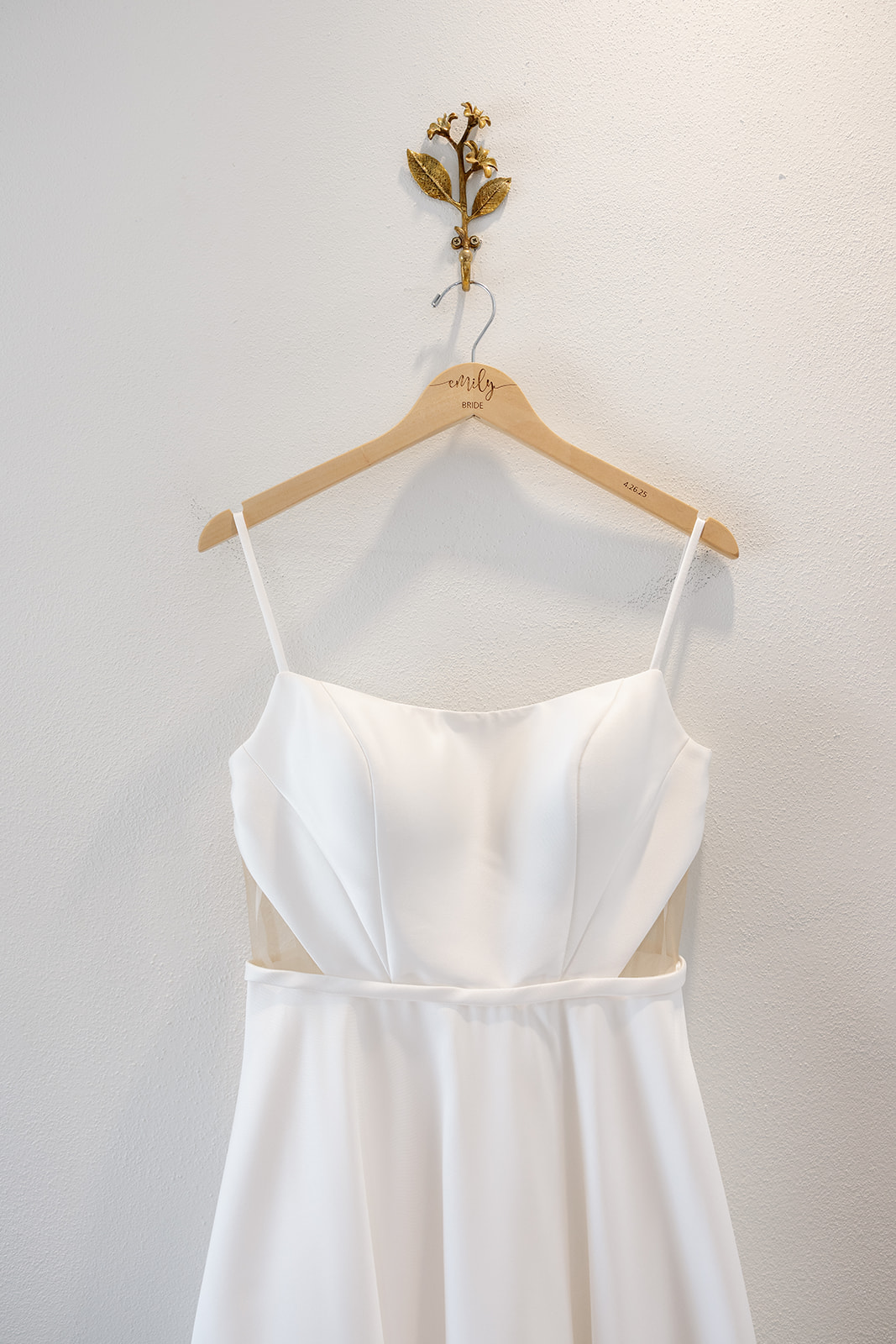 Simple white wedding gown with spaghetti straps and illusion side panels hanging on a floral hook.