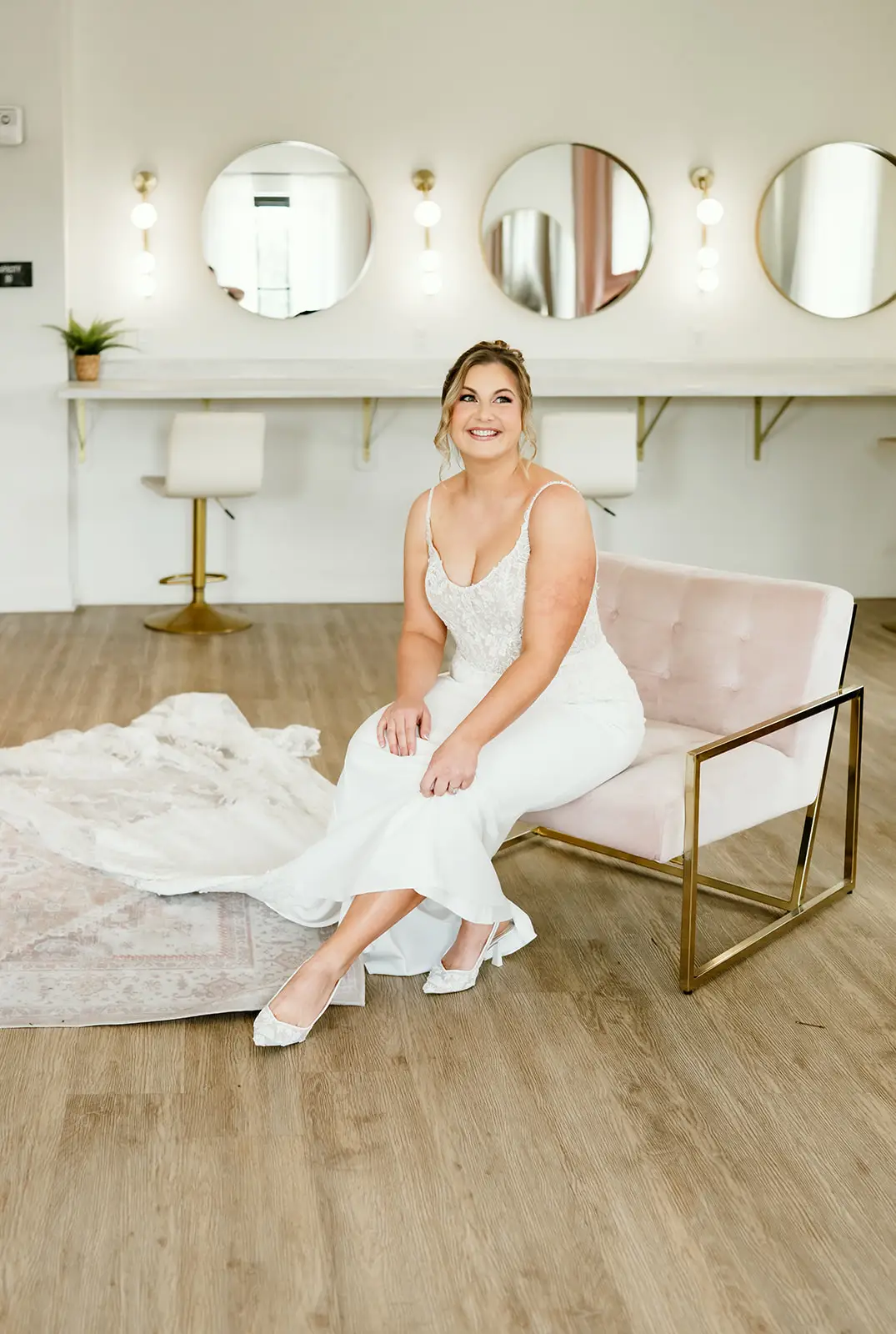 Bride smiles while seated in modern bridal suite at 10 South with vanity mirrors, pink velvet chair, and gown nearby