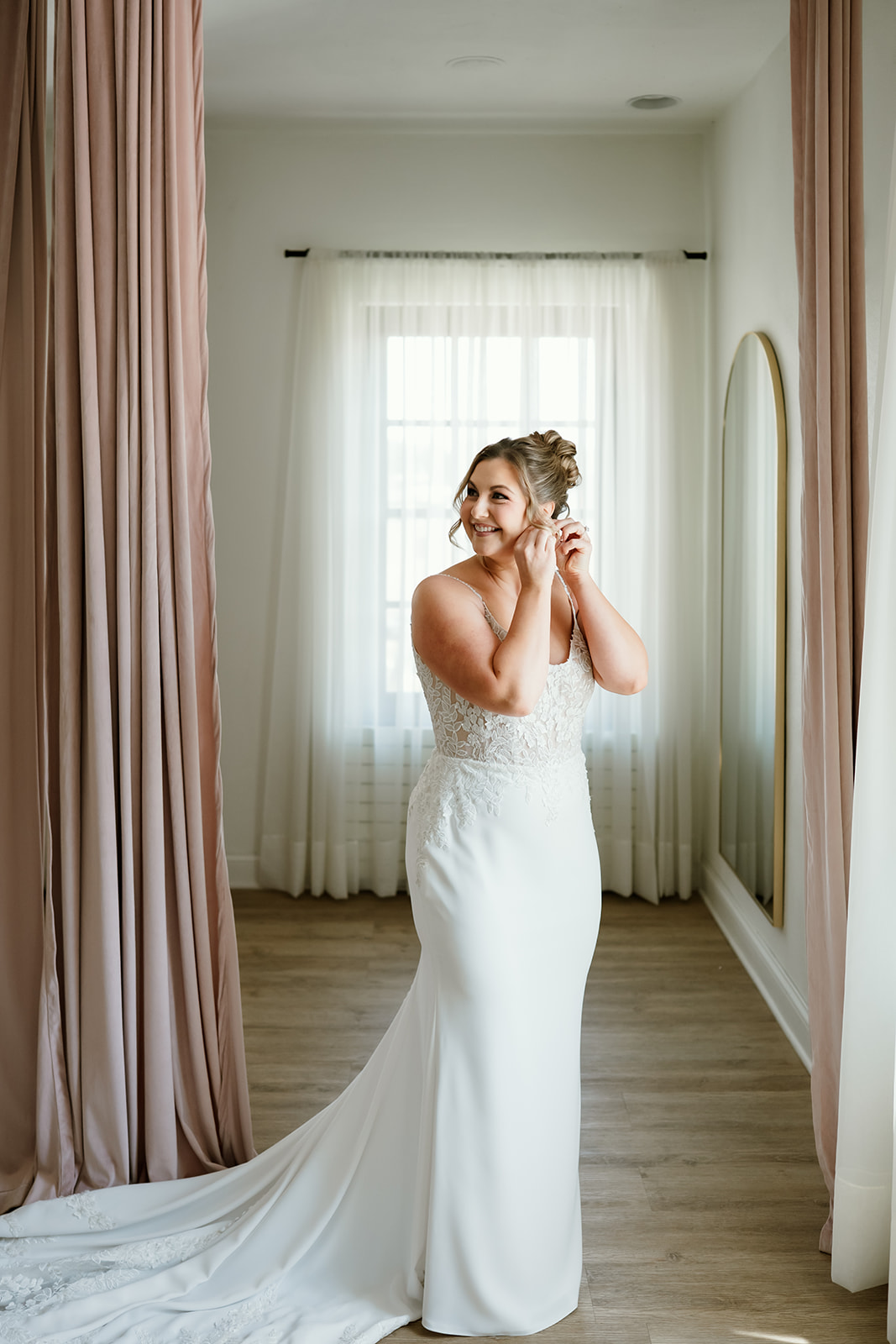 Bride adjusts earring while standing in a bridal dressing suite with blush curtains and natural light.
