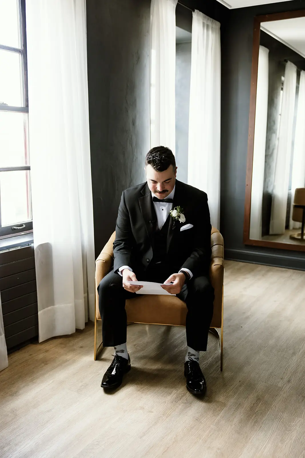 Groom in black tuxedo sits reading a letter in the groom’s lounge at 10 South in Janesville, WI